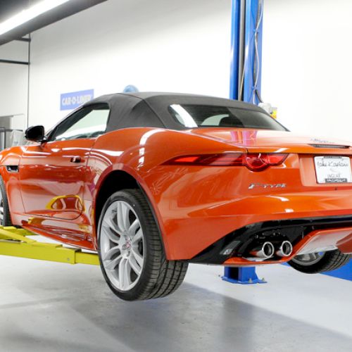 An orange convertible car is lifted on a mechanic's hydraulic lift, with a focus on the rear and exhaust, in an indoor garage space.