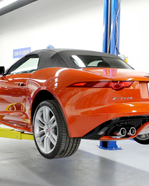 An orange convertible car is lifted on a mechanic's hydraulic lift, with a focus on the rear and exhaust, in an indoor garage space.
