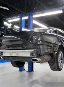 Two cars are lifted on hydraulic lifts in a well-lit auto repair shop, with one missing a rear bumper.