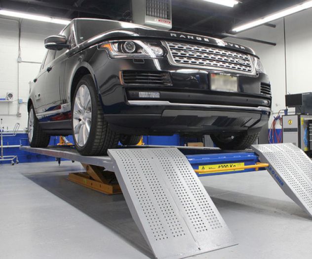 A black SUV is on a vehicle lift inside a garage or service center, with ramps leading up to the platform.