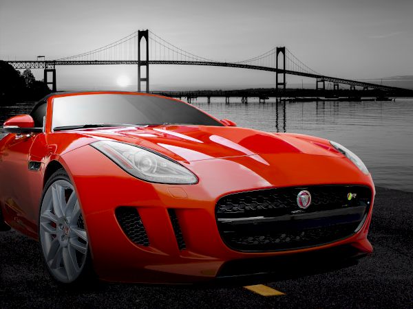 A red sports car is parked on a road, with a bridge and a body of water in the background, under a grayscale sky.