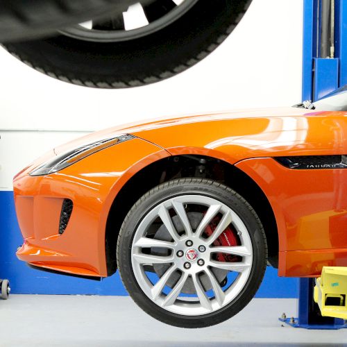 An orange sports car is elevated on a vehicle lift in a workshop, with a blue Eurovac system nearby and a tire visible above.