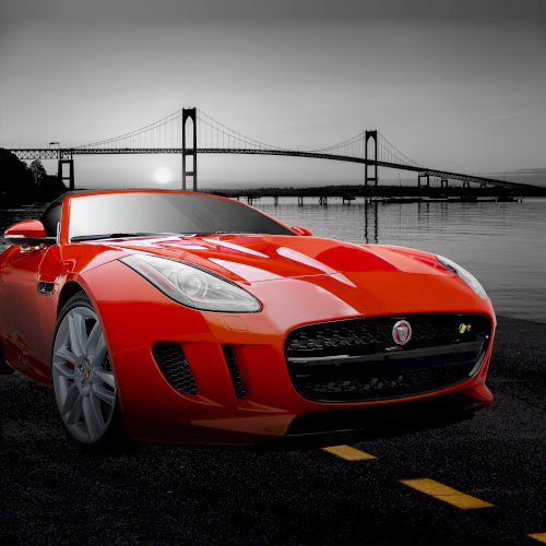 A red sports car is parked on a road near a waterfront, with a suspension bridge in the background under a gray sky.